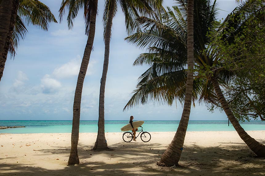 Woman cycling along the beach with a surfboard in the Maldives